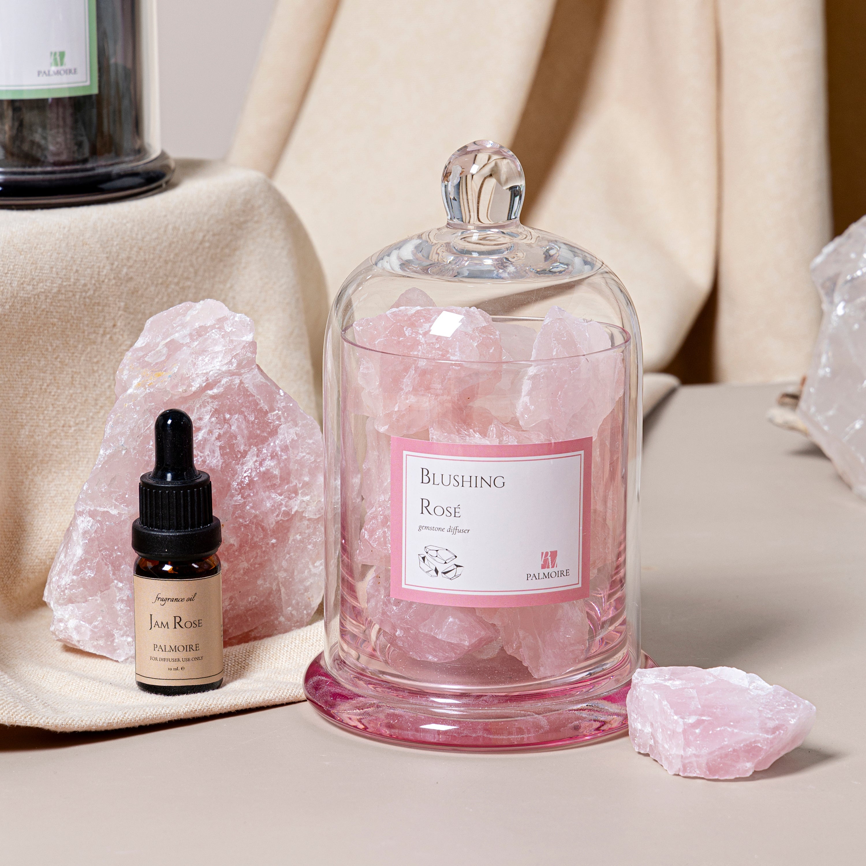 Glass container labeled 'Blushing Rose' with pink crystals, a small bottle, and rose quartz on a neutral background.